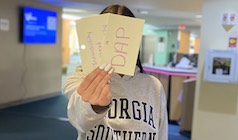 A student holds a notecard reading "DAP"; she stands in a student lounge area and is wearing a "Georgia Southern" sweatshirt.