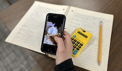 A student scrolls on a mobile phone while a notebook, calculator, and pencil sit in the background.