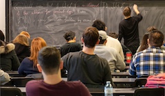 Students sit in a classroom watching their instructor write on a chalk board.